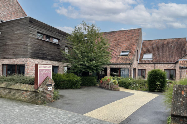 Woonzorgcentrum Anemoon in Puurs, gezien vanaf de straat. Het gebouw is goed zichtbaar, met bakstenen gevels en houten accenten. De naam van het woonzorgcentrum staat duidelijk op een bord, en een pad leidt naar de ingang.