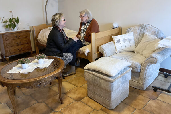 Een bewoner van woonzorgcentrum Anemoon zit op haar bed in haar kamer, terwijl iemand naast haar bukt en haar aandacht geeft. De kamer is ingericht met een tafeltje, een houten nachtkastje en een eenpersoonssofa.