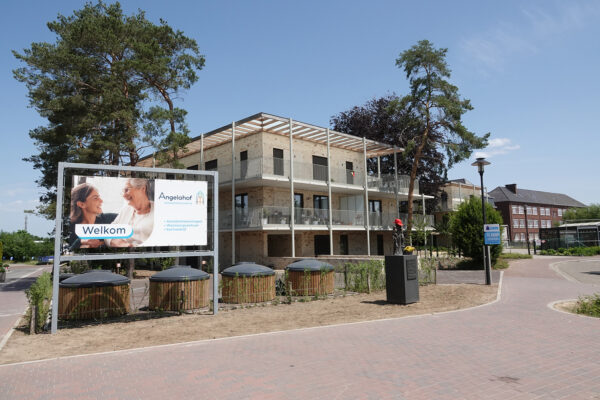 Een pad dat het gebouw van het woonzorgcentrum omringt, met op de achtergrond het gebouw van gele baksteen. Op de voorgrond is een plakkaat zichtbaar die iedereen verwelkomt die naar het woonzorgcentrum komt.