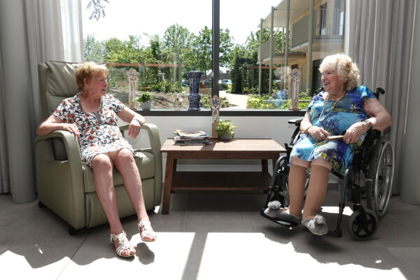 Twee bewoners van woonzorgcentrum Angelahof zitten samen naast een groot raam, waardoor veel natuurlijk licht binnenvalt, en voeren een gezellig gesprek.