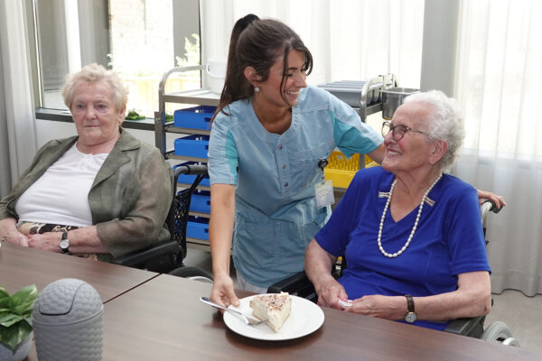Een medewerker van woonzorgcentrum Angelahof serveert een taart als dessert aan een bewoner die samen met een andere bewoner aan tafel zit tijdens de maaltijdsservice.