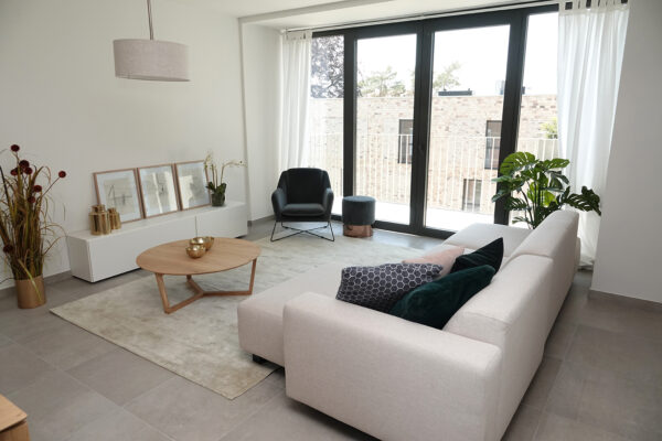 De zitkamer van een assistentiewoning in Angelahof, ingericht met een grote witte sofa en een zwarte fauteuil. Vanuit de zitkamer is het terras bereikbaar via grote glazen dubbele deuren, die veel natuurlijk licht binnenlaten.
