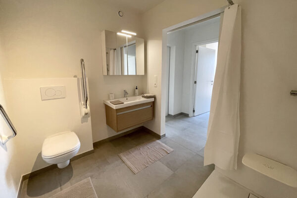 Een ruime, moderne badkamer in een assistentiewoning van Angelahof, uitgerust met alle benodigde hulpvoorzieningen. De badkamer heeft een witte wastafel met daaronder opbergruimte. De douche is drempelloos en gemakkelijk toegankelijk, en is voorzien van een zitbankje.