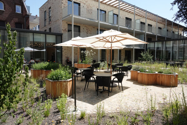 Het grote terras van woonzorgcentrum Angelahof, waar bewoners onder de zon kunnen samenzitten. Grote parasols bieden schaduw voor wie dat wenst. Het terras is omringd door planten en een groene omgeving en is direct bereikbaar vanuit het gebouw, dat zichtbaar is op de achtergrond.