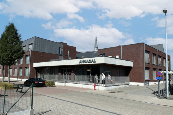De ingang van woonzorgcentrum Annadal, gezien vanaf de straat. Boven de ingang is de naam van het woonzorgcentrum duidelijk zichtbaar. Het gebouw heeft gevels van donkerrode bakstenen. Drie mensen betreden het woonzorgcentrum via een helling, die het voor iedereen gemakkelijk toegankelijk maakt, ook voor rolstoelgebruikers.