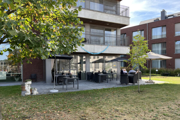 Foto genomen vanuit de tuin van woonzorgcentrum Annadal in Retie, met zicht op het gemeenschappelijke terras en de verschillende gebouwen van het woonzorgcentrum. Boven het terras zijn de hoger gelegen slaapkamers zichtbaar, die beschikken over een privéterras.