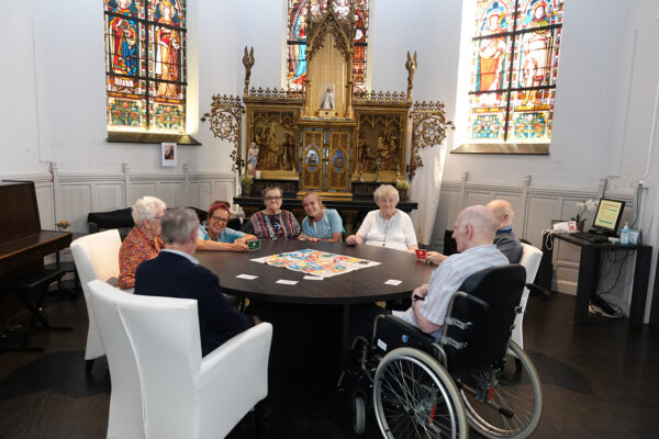Zeven bewoners en twee medewerkers zitten samen aan een ronde tafel in woonzorgcentrum Annadal in Retie, waar ze een gezelschapsspel spelen. De ruimte is voorzien van glas-in-loodramen, waarop verschillende mensen zijn afgebeeld.