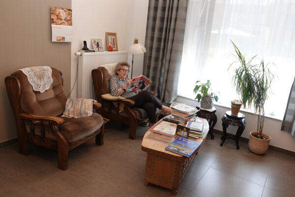 Een kamer in woonzorgcentrum Annadal, waar een bewoonster op een van haar twee bruine fauteuils zit en een tijdschrift leest. Op de houten bijzettafel in het midden van de kamer liggen meerdere tijdschriften en boeken. Er is een groot raam dat veel natuurlijk licht naar binnen laat stromen.