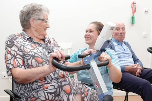 Twee bewoners en een medewerker zitten samen in de kinesitherapieruimte van woonzorgcentrum Annadal, waarbij de medewerker een vrouwelijke bewoner helpt met haar trapoefeningen.