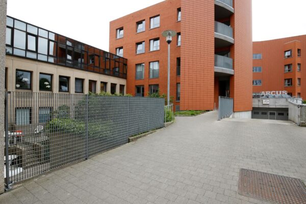 De voorgevel van het neuropsychologisch woonzorgcentrum Arcus in Brussel is zichtbaar, met de ingang van het gebouw van rode bakstenen. Ook is de garagedeur te zien, die toegang biedt tot de ondergrondse garage. De naam van het woonzorgcentrum is duidelijk zichtbaar aan de buitenkant. Links in de foto, achter een hek, is het terras van het woonzorgcentrum te zien.