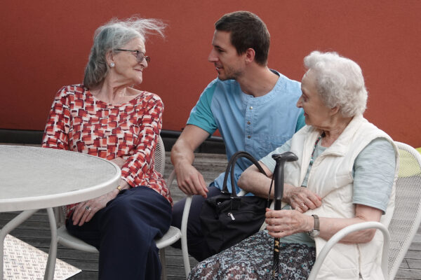 Twee bewoners van het neuropsychologisch woonzorgcentrum Arcus in Brussel zitten samen op het terras, in een gezellig gesprek met een medewerker die naast hen gebukt is, zodat ze elkaar op ooghoogte kunnen aankijken.