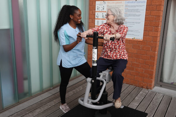 Een bewoner van woonzorgcentrum Arcus doet fietsoefeningen op een apparaat op het terras van het woonzorgcentrum, terwijl een medewerkster haar begeleidt. Op de achtergrond is een plakkaat zichtbaar waarop in zowel het Nederlands als het Frans wordt uitgelegd hoe het apparaat correct gebruikt moet worden.