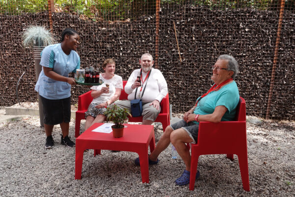 Drie bewoners van neuropsychologisch woonzorgcentrum Arcus zitten op rode stoelen in de tuin, gezellig met elkaar in gesprek. Een medewerker brengt hen een drankje en een ijsje.