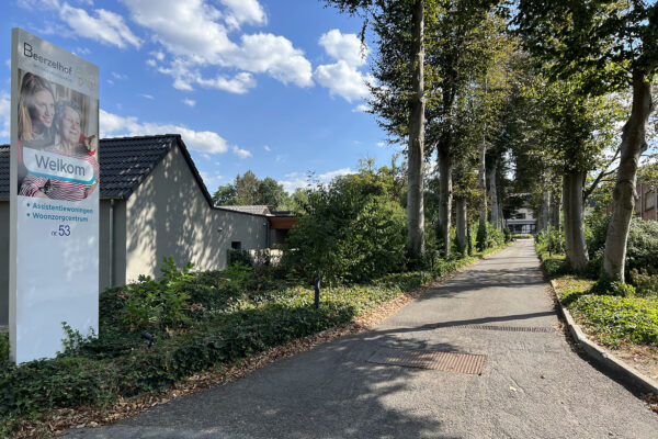 Het pad dat met de auto genomen kan worden om direct bij woonzorgcentrum Beerzelhof te komen. Een signalisatie is zichtbaar, met de naam en het logo van het woonzorgcentrum. Deze verwelkomt bezoekers en informeert hen dat zowel een woonzorgcentrum als assistentiewoningen op de campus aanwezig zijn.
