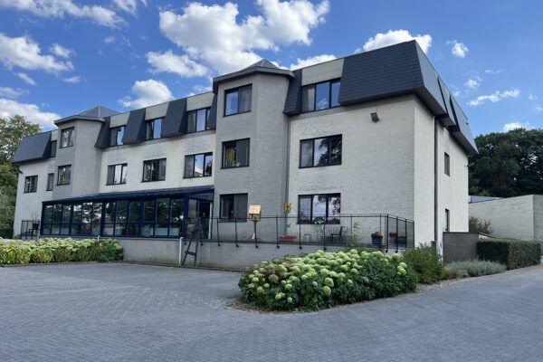 De voorgevel van woonzorgcentrum Beerzelhof, gebouwd uit witte en grijze bakstenen, gezien vanuit de parking. Ook is het terras zichtbaar, dat bereikbaar is vanuit de serre van het gebouw.