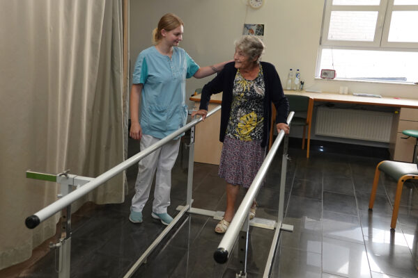 Een bewoner voert wandeloefeningen uit in de kinesitherapieruimte van woonzorgcentrum Beerzelhof. De medewerkster staat naast haar, biedt steun waar nodig en assisteert haar tijdens de oefeningen.