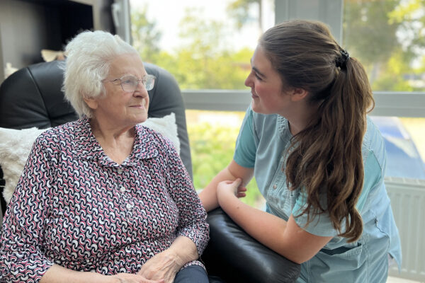 Een bewoner van woonzorgcentrum Beerzelhof zit in het salon op een zwarte fauteuil, terwijl een medewerkster met haar in gesprek is en aandachtig luistert.