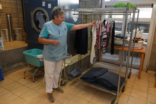 Het waslokaal van woonzorgcentrum Beerzelhof, waar een medewerkster de net gewassen kleren van de bewoners ophangt. Op de achtergrond draait de wasmachine, terwijl andere kleding klaarstaat om gewassen te worden.