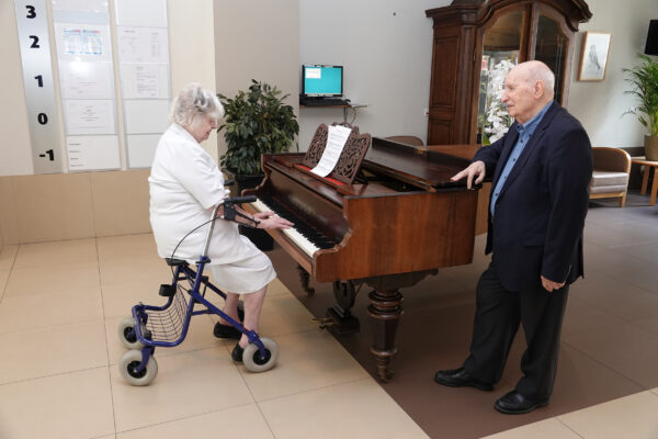 Een piano in de inkomhal van woonzorgcentrum Bellevue. Een bewoner heeft plaatsgenomen achter de piano en speelt muziek, terwijl een andere bewoner naast de piano staat en geniet van de melodieën.