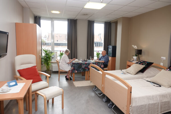 Een dubbele kamer in woonzorgcentrum Bellevue, waar een man samen met zijn vrouw woont. Ze zitten gezellig samen te eten. De kamer is ingericht met een stoel en voetbankje, een kast, een houten tafel, een televisie en twee bedden, elk met een nachtkastje.