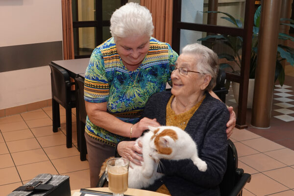 Twee bewoners van woonzorgcentrum Borsbeekhof bevinden zich in de bar van het woonzorgcentrum. Een van hen heeft haar kat mee en geniet van een kopje koffie, terwijl de andere bewoner dichterbij komt om de kat te bewonderen.
