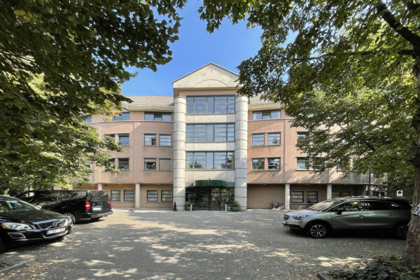 De ingang van woonzorgcentrum Borsbeekhof, gezien vanaf de buitenkant. Bezoekers kunnen hun auto parkeren op de parkeerplaats voor de ingang. Het gebouw, met zijn ronde vorm, is duidelijk zichtbaar. De grote pilaren markeren duidelijk de locatie van de voordeuren.