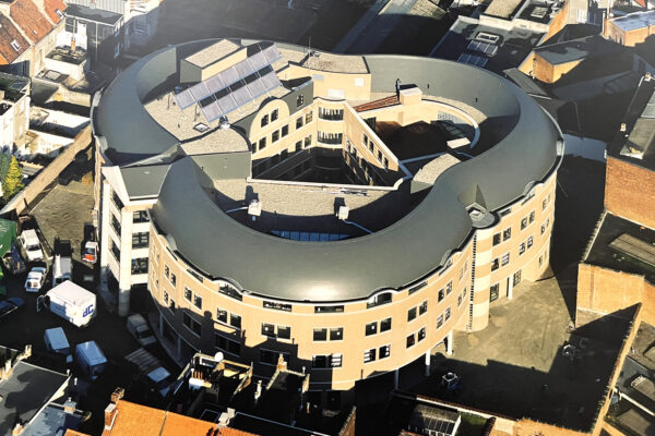 Woonzorgcentrum Borsbeekhof vanuit de lucht gezien. De unieke vorm van het gebouw is duidelijk zichtbaar, evenals de zonnepanelen die op het dak zijn geplaatst. In het midden van het gebouw bevindt zich een opening waar het terras is gelegen.