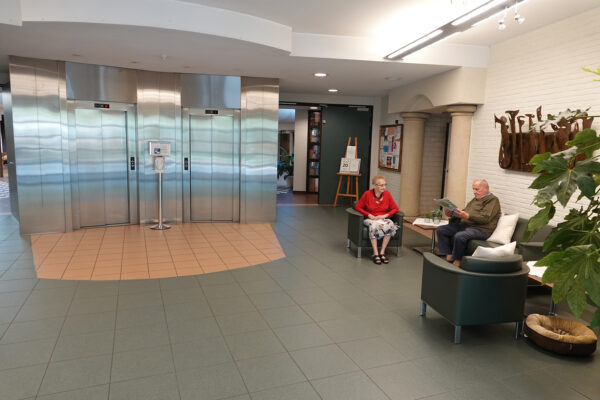 De inkomhal van woonzorgcentrum Borsbeekhof in Borgerhout. In de hal staan comfortabele sofa’s waar bewoners kunnen zitten. Twee bewoners rusten daar, waarvan één de krant van de dag leest. De inkomhal bevat ook de liften, die bewoners naar hun kamers kunnen brengen.