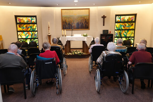 De kapel van woonzorgcentrum Borsbeekhof, waar de bewoners samen zitten tijdens de mis. De ruimte is voorzien van prachtige, gedecoreerde gekleurde ramen en is ingericht met een kruis en een groot schilderij dat een groep mensen toont die samen een zonsopgang aanschouwen.