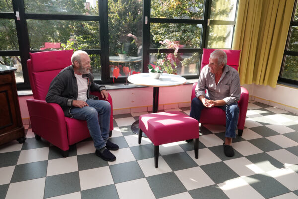 Het salon van woonzorgcentrum Borsbeekhof, ingericht met grote rode fauteuils, een rood voetbankje en een ronde bijzettafel. Het salon wordt omringd door grote ramen, waardoor er veel zonlicht binnenkomt. De beplanting aan de buitenkant is ook goed zichtbaar van binnen. Twee bewoners zitten in de rode fauteuils en voeren een gezellig gesprek met elkaar.