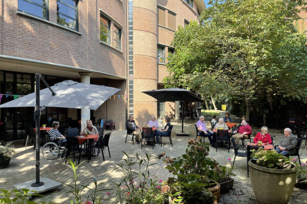 Het terras van woonzorgcentrum Borsbeekhof in Borgerhout, waar meerdere bewoners en hun familieleden samen rond verschillende tafels zitten. Het terras is omgeven door grote bomen en weelderige planten. Ook is de bakstenen gevel van het gebouw zichtbaar, die direct vanuit het terras toegankelijk is.