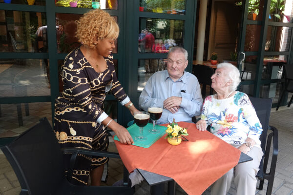 Het terras van woonzorgcentrum Borsbeekhof in Borgerhout. Twee bewoners zitten aan een tafel en krijgen beiden een drankje aangeboden door een vrouwelijke vrijwilliger.