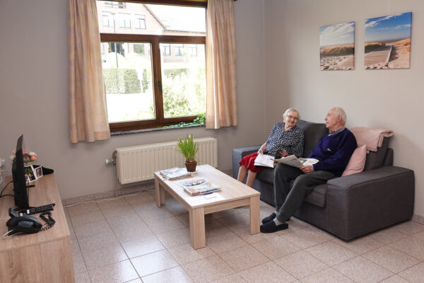 Twee bewoners van woonzorgcentrum De Vlaamse Ardennen zitten samen op een sofa in hun kamer, voor de televisie.