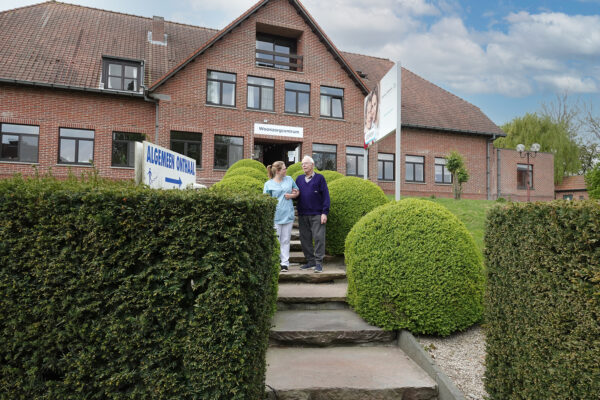 Trappen naar de inkom van woonzorgcentrum De Vlaamse Ardennen. Een bewoner is een wandeling aan het doen met een medewerker. De voorgevel van het gebouw is zichtbaar.