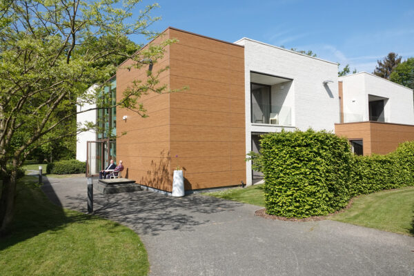 Een complex met meerdere assistentiewoningen, waarvan de gevels gedeeltelijk uit witte bakstenen en gedeeltelijk uit hout bestaan. Twee bewoners zitten aan de ingang van het gebouw aan een klein wit tafeltje en voeren een gesprek.