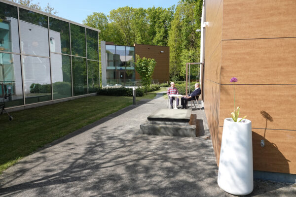 Twee bewoners zitten op het terras van hun assistentiewoning op de begane grond. Het terras biedt directe toegang tot de tuin en de andere gebouwen van de campus.