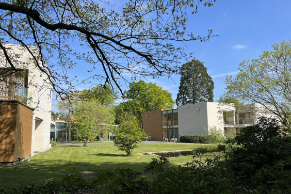 Een aantal gebouwen met een gelijkaardige architectuur is gedeeltelijk zichtbaar, waarbij een gebouw met een glazen gevel tussen hen uitsteekt. De gebouwen zijn omringd door een overvloed aan groen, met veel planten en bomen.