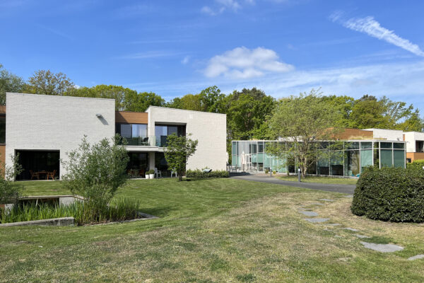 De buitengevel van een assistentiewoning in Ter Bleuk, volledig opgebouwd uit witte bakstenen met een houten accent bovenaan, is aan de linkerkant van de foto zichtbaar. Aan de rechterkant is het administratieve gebouw te zien, dat volledig uit glas bestaat. Op de voorgrond ligt de tuin, terwijl op de achtergrond een weelderig groen bomenlandschap te zien is.