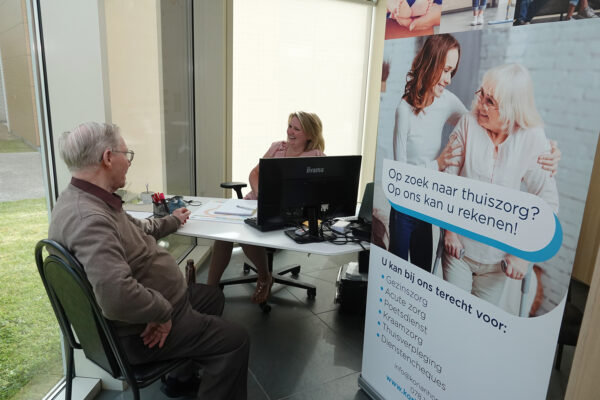 Het bureau van het assistentiewoningencomplex, waar een medewerkster een nieuwe bewoner verwelkomt. Op een plakkaat is te lezen dat er ook gezinszorg beschikbaar is via het Korian Home Care bureau.