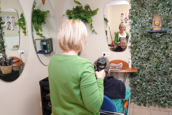 De kapsalon in de assistentiewoningen Ter Bleuk in Rijmenam. De kapper is het net gewassen en geknipte haar van een bewoner aan het drogen, terwijl de bewoner met blijdschap haar spiegelbeeld bewondert. Aan de muren hangen drie spiegels, en één muur is versierd met kunstplanten.