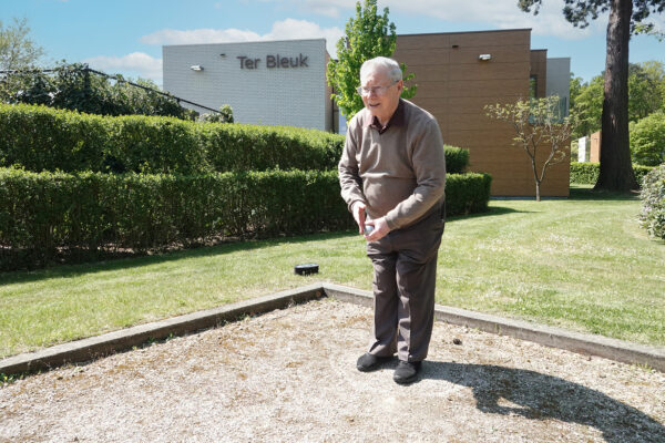 Een bewoner van de assistentiewoningen Ter Bleuk in Rijmenam speelt alleen een potje petanque. Op de achtergrond is een gebouw zichtbaar, met de naam van het complex duidelijk vermeld.