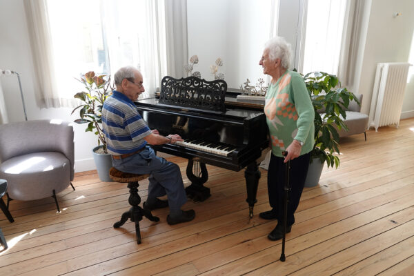 Twee bewoners van woonzorgcentrum Angelahof bevinden zich samen in een ontspanningsruimte. Eén van de bewoners zit naast een piano en speelt erop, terwijl de andere rustig naast hem staat en geniet van de muziek.