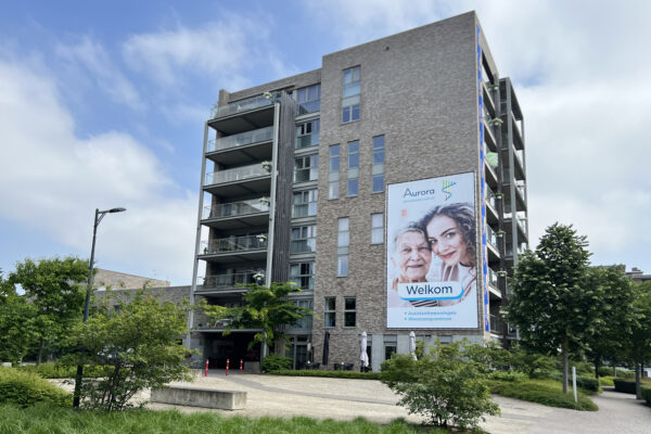 Het gebouw van woonzorgcentrum Aurora, gezien vanuit de buitenkant, is omgeven door een groene omgeving met veel bomen. Op de gevel hangt een groot bord waarop bezoekers welkom worden geheten. Het bord toont de naam van het woonzorgcentrum, inclusief het logo, en vermeldt dat er zowel assistentiewoningen als een woonzorgcentrum beschikbaar zijn.