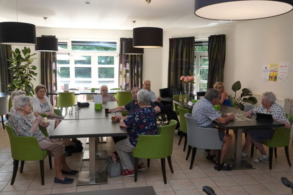 De brasserie van woonzorgcentrum Beerzelhof. Aan een tafel zitten zes bewoners gezellig te drinken en te praten. Aan een andere tafel spelen twee bewoners een kaartspel, terwijl een andere bewoner samen met een familielid van een drankje geniet.