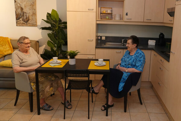 Een bewoner van een assistentiewoning in Beerzelhof zit samen met haar bezoek aan de eettafel, naast de keuken. Ze zitten gezellig aan de tafel, drinken koffie en voeren een ontspannen gesprek.