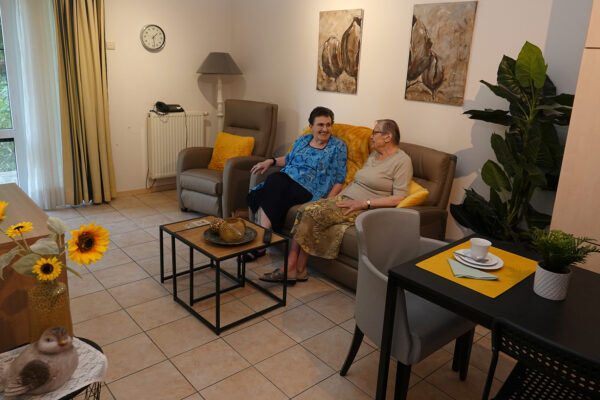 De woonkamer van een assistentiewoning in Beerzelhof, ingericht met een bruine sofa en een bijpassende fauteuil. De bewoner zit samen met haar bezoek op de sofa, terwijl ze gezellig met elkaar in gesprek zijn. Twee moderne bijzettafels staan in de kamer, en aan de muur hangen twee schilderijen die de ruimte verder sieren.