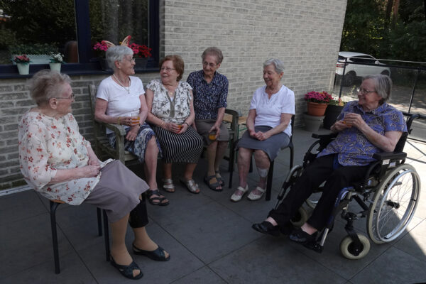 Zes bewoners zitten samen op het terras van woonzorgcentrum Beerzelhof. Terwijl ze van het frisse buitenlucht genieten, drinken ze een drankje en praten ze gezellig met elkaar.