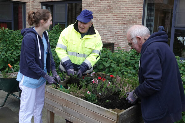 Twee bewoners van woonzorgcentrum Berckenbosch zijn samen met een medewerkster aan het tuinieren. Alle drie dragen tuinhandschoenen. Eén van de bewoners draagt een fluojas en een pet en is in gesprek met de medewerkster.