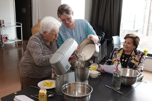 De animatiemedewerkster van woonzorgcentrum Berckenbosch in Heusden bereidt samen met twee bewoners wafels. Eén bewoner zit aan de tafel en leest het recept voor, terwijl de andere twee bewoners bloem in een kom aan het doen zijn.