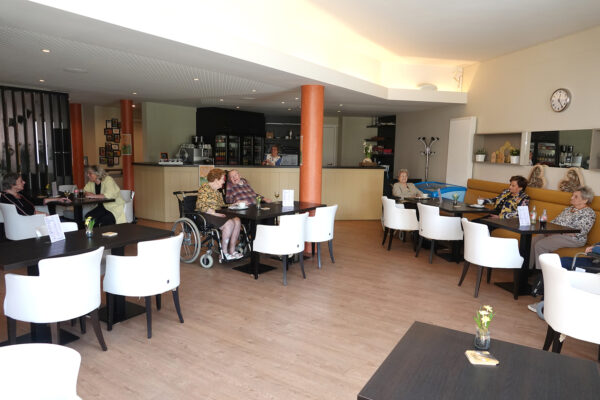 De brasserie van woonzorgcentrum Berckenbosch, waar bewoners terecht kunnen voor een hapje of een drankje. Een vrijwilliger bedient de bar. Acht bewoners zitten aan tafels, genietend van een drankje. Sommigen zitten alleen, terwijl anderen met zijn tweeën aan een tafel zitten en met elkaar in gesprek zijn.